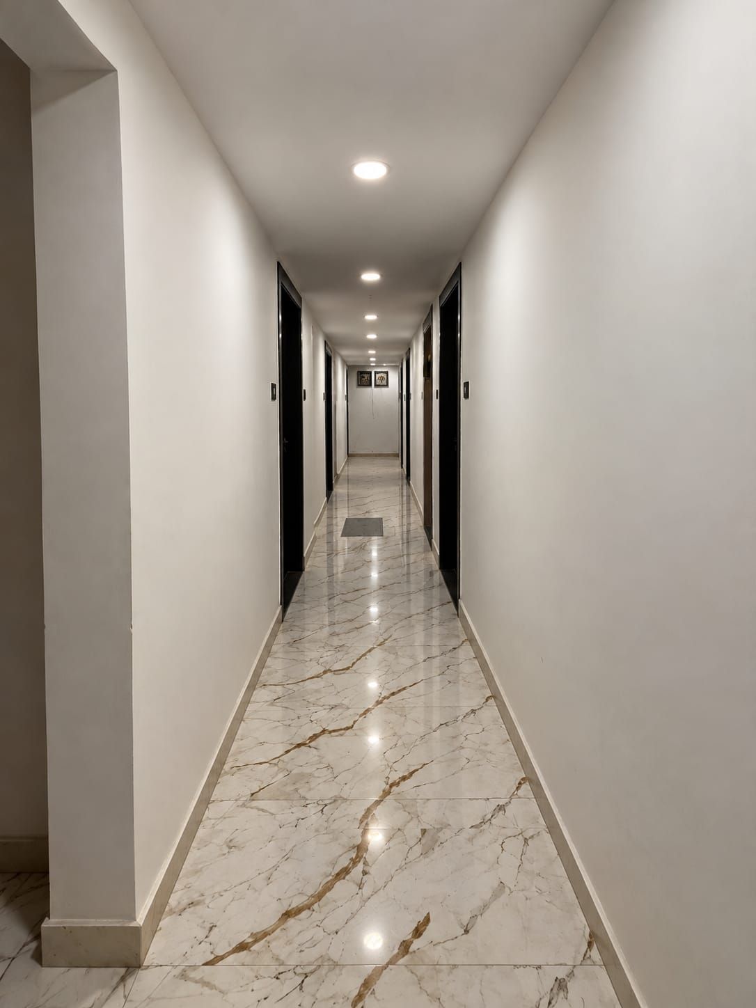 Un largo pasillo estrecho con paredes blancas y suelo de mármol, que conduce a una puerta al final.