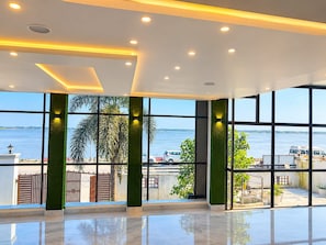 A spacious interior with large windows offering a view of the water and palm trees. - Sapphire The Ganga View (Prayagraj)