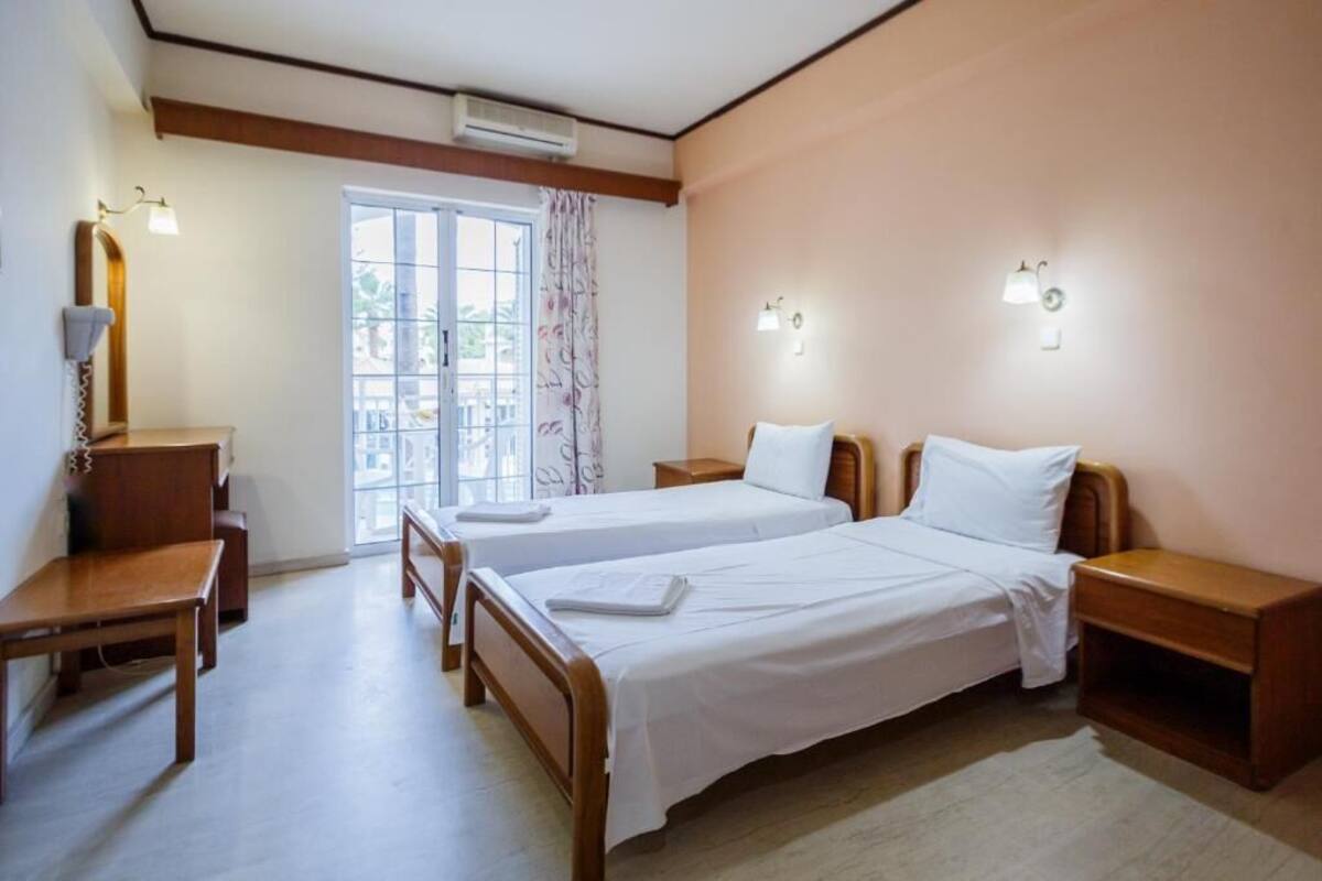 A hotel room with two single beds, a wooden desk, a chair, a small table, and a window with curtains.