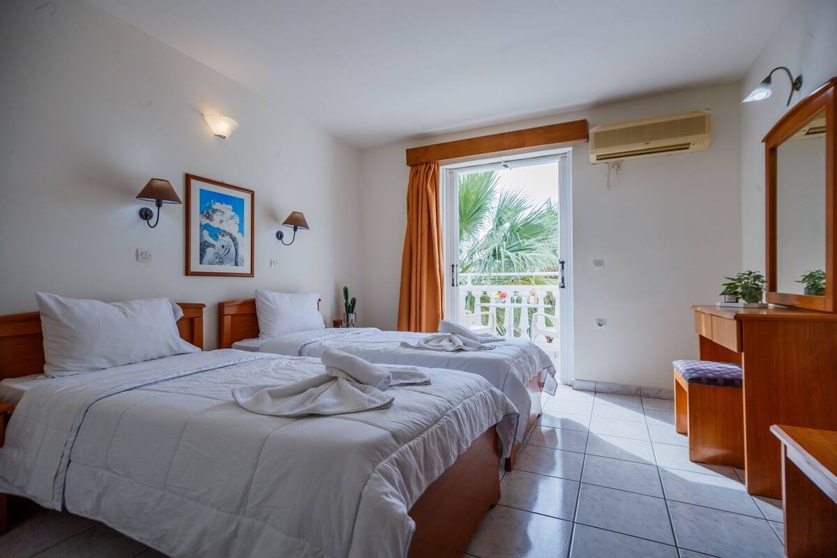 A hotel room with two beds, a wooden dresser, a window with curtains, an air conditioning unit, and a framed picture on the wall.