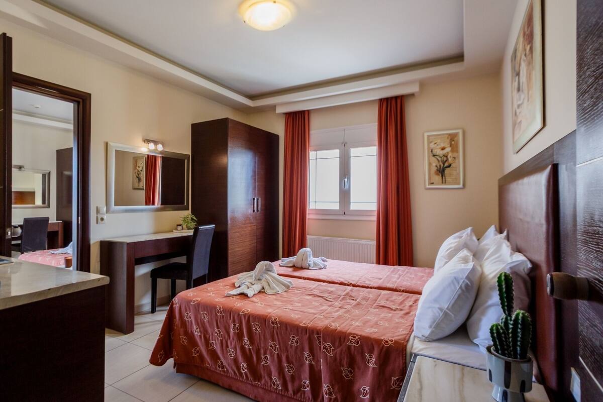 A hotel room with a bed, a desk, a chair, a mirror, a window with curtains, and a cactus plant.