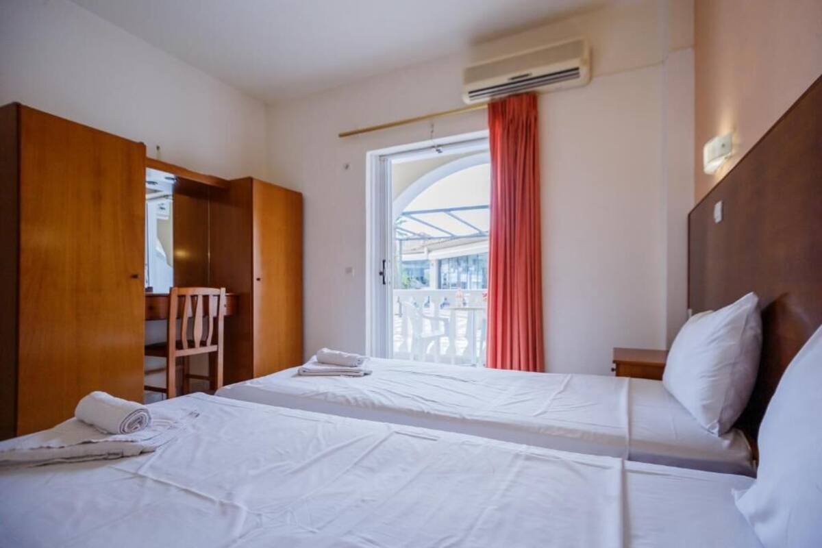 A hotel room with two beds, a wooden wardrobe, a chair, and a balcony with a view.
