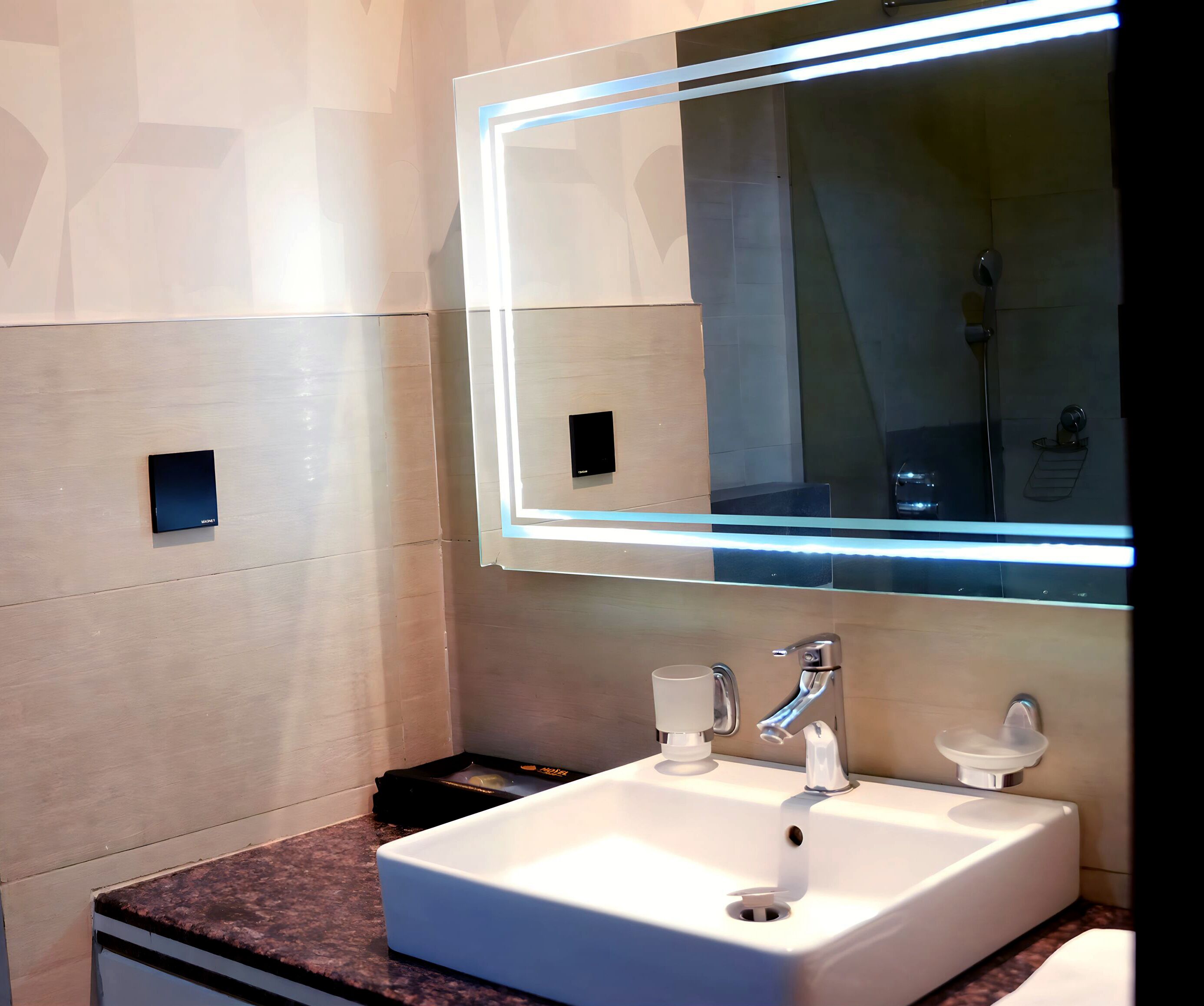 Ein modernes Badezimmer mit einem weißen Waschbecken, zwei Wasserhähnen, einem Spiegel mit Lichtleiste und einer kleinen Mülleimer.