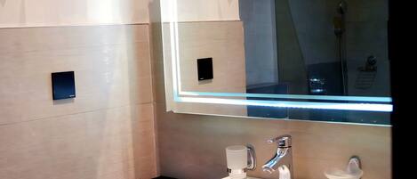 Un baño moderno con un lavamanos blanco, dos llaves, un espejo con iluminación y un pequeño cesto de basura.