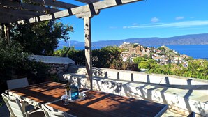 An outdoor patio with a wooden table and chairs, a stone wall, and a view of a coastal village.