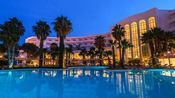 Een hotel met een groot zwembad, palmbomen en een gebouw met rondboogvensters.