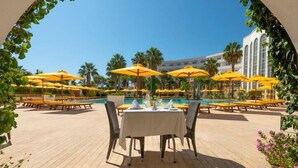 Een eetgedeelte bij het zwembad met tafels en stoelen, gele parasols en vrij uitzicht op het zwembad en palmbomen.