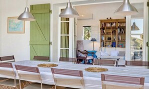 A dining area with a table set for four, a bookshelf, and a chair with a blue cushion. - CASA-Stella Maris villa avec vue sur la mer (Cavalaire-sur-Mer)