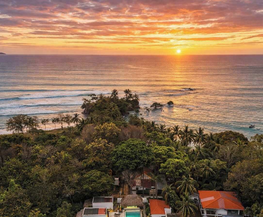 A coastal view with a sunset, a beach, and a cluster of houses surrounded by dense vegetation.