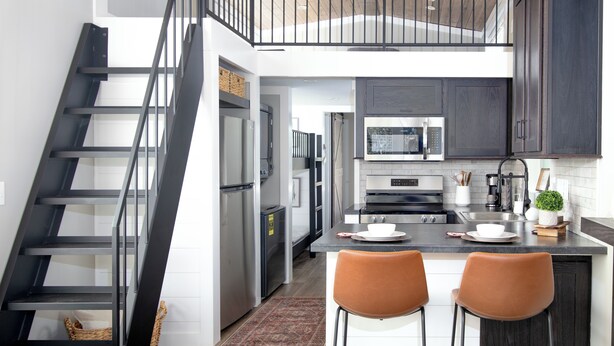 A modern kitchen with dark cabinetry, stainless steel appliances, a central island with seating, and a wooden staircase. - Mountain Tiny Home in Blue Ridge Mountains (Copperhill)