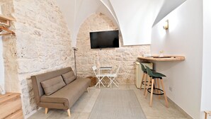 Un accogliente soggiorno con parete in pietra, una TV a schermo piatto posizionata sopra un tavolo di legno con sedie, un divano e un piccolo sgabello in legno.
