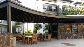 An outdoor patio area with wooden tables and chairs, a stone wall, and a covered seating area. - Furnished Apartment in Altea Condominiums, Puerto Vallarta, Jalisco. (Puerto Vallarta)