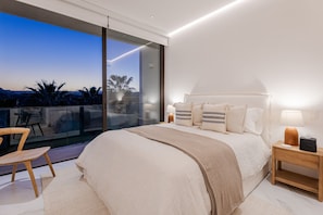 A modern bedroom with a large bed, bedside lamps, and a view of palm trees through a sliding glass door. - Cora Sunset: Brand New Oceanview 3BR Near Golf, Pool & Swimmable/Surfable Beach (San José del Cabo)