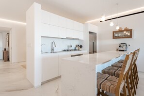 A modern kitchen with white cabinetry, a large island with bar stools, and a built-in oven. - Cora Sunset: Brand New Oceanview 3BR Near Golf, Pool & Swimmable/Surfable Beach (San José del Cabo)