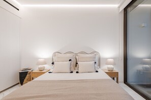 A minimalist bedroom with a large bed, two bedside tables with lamps, and a view through a glass door. - Cora Sunset: Brand New Oceanview 3BR Near Golf, Pool & Swimmable/Surfable Beach (San José del Cabo)