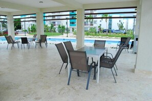 A covered outdoor dining area with a pool view, featuring a table and chairs, and a view of a residential building in the background. - Amor - 1 BR apartment (Punta Cana)