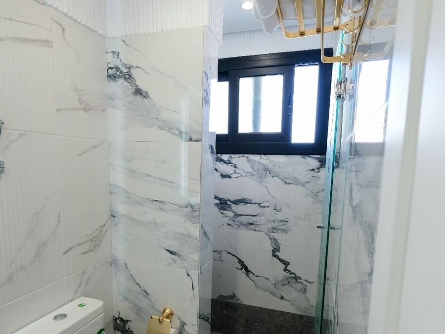 A bathroom with a toilet, marble walls, a shower with glass doors, and a window.