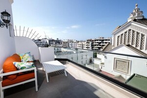 Un balcón con un sofá blanco y una pequeña mesa, con vistas a un paisaje urbano de edificios blancos y una mezquita de diseño intrincado.