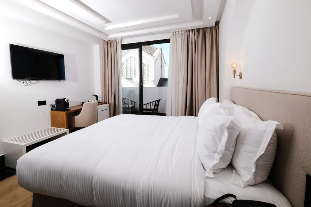 A hotel room with a large bed, a desk with a chair, a flat-screen TV mounted on the wall, and a balcony with curtains.