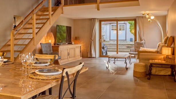 Amplia sala de estar con una escalera de madera, una mesa de comedor puesta con platos y vasos, un sofá y vistas al exterior.
