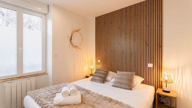 Een slaapkamer met een bed, een wand van houten latten, een raam en een nachtkastje met een lamp.