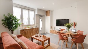 Een moderne woonkamer met een bank, een eettafel met stoelen, een aan de muur bevestigde televisie en een groot raam.
