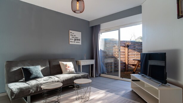 Una sala de estar moderna con un sofá gris, una mesita redonda pequeña, un televisor sobre un mueble blanco y un amplio ventanal corredero que da a un espacio exterior.