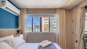 Une chambre d’hôtel avec un lit, une fenêtre avec des rideaux, un climatiseur fixé au mur et une vue sur les bâtiments environnants.