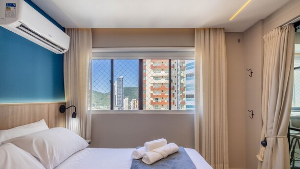 Camera d'albergo con un letto, una finestra con tende e condizionatore d'aria a muro, con vista sugli edifici.