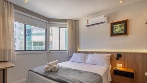 Une chambre d’hôtel comprenant un lit, une table de chevet, un appareil de climatisation, une fenêtre avec des rideaux et un tableau encadré au mur.