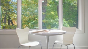 A room with a round table and two white chairs, a hanging light fixture, and a view of green trees through large windows. - The Private Oasis • 22 min to downtown (Indianapolis)