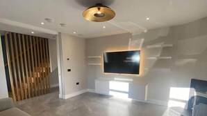 A modern living room with a large flat-screen TV mounted on a wall, a grey sofa, and a wooden slatted partition. - Dingle Doorstep - Town Centre (Dingle)