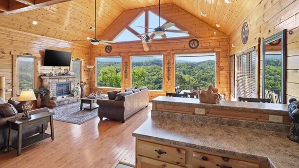 A log cabin interior with a fireplace, a sofa, a coffee table, and a large window offering a view of the forest. - NEW! Luxury Cabin w/Hot Tub & Stunning Mntn Views! (Sevierville)