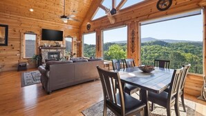 A wooden cabin interior with a dining table, chairs, a sofa, a fireplace, and large windows offering a forest view. - NEW! Luxury Cabin w/Hot Tub & Stunning Mntn Views! (Sevierville)