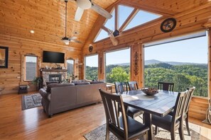 Ein Holzblockbau-Innenraum mit Esstisch, Stühlen, einer Couch, einem Kamin und großen Fenstern mit Blick auf einen Wald.
