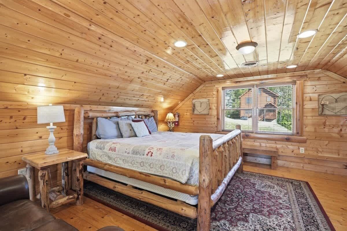 A wooden cabin bedroom with a bed, a nightstand, a lamp, and a window with a view of a house.