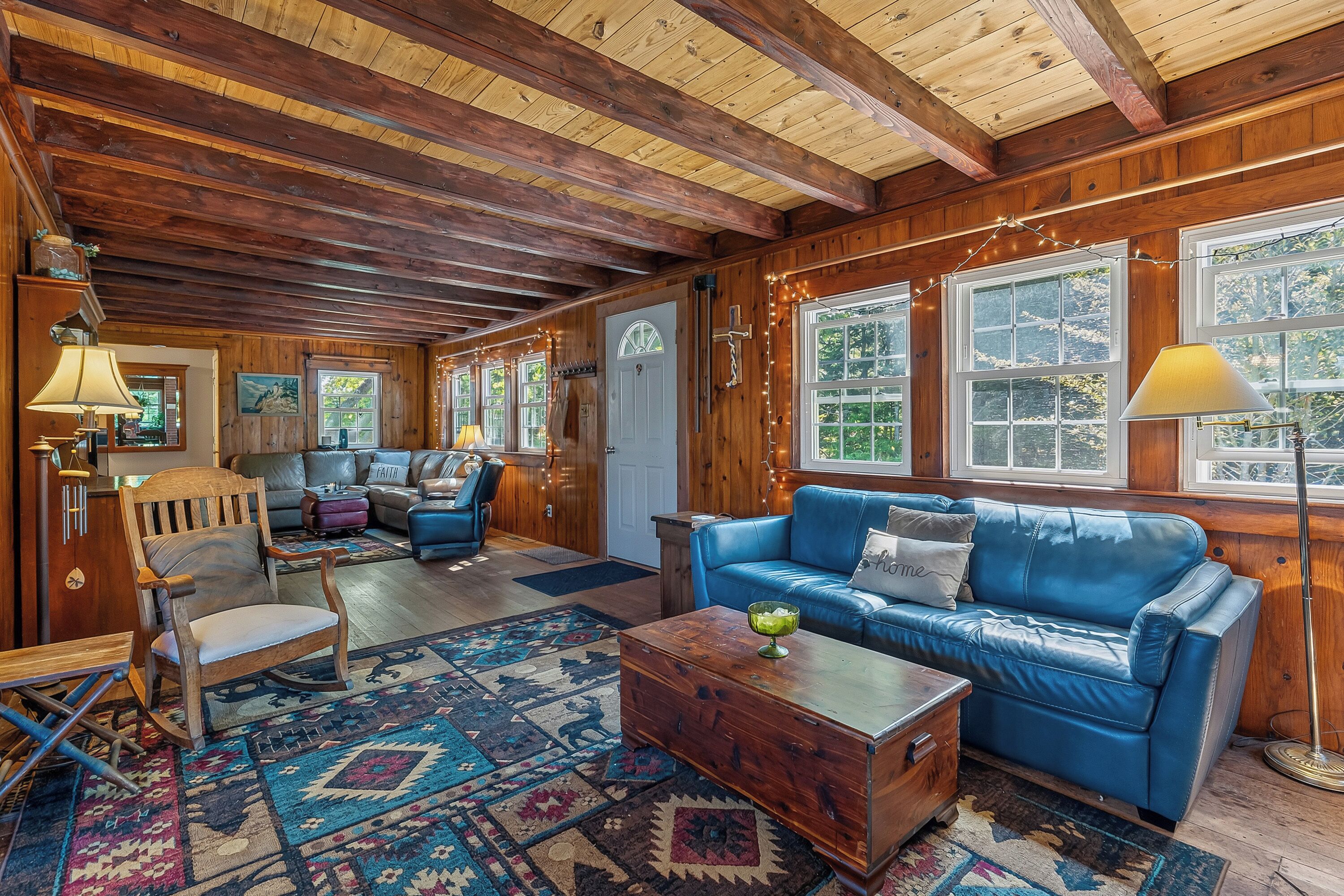 A cozy cabin interior with a wooden ceiling, a blue leather sofa, a wooden chest, and a patterned rug.