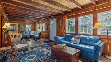 A cozy cabin interior with a wooden ceiling, a blue leather sofa, a wooden chest, and a patterned rug.