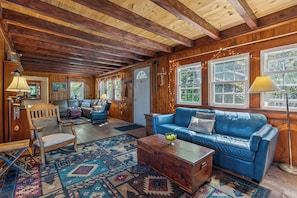 A cozy cabin interior with a wooden ceiling, a blue leather sofa, a wooden chest, and a patterned rug. - 5-bedroom Lodge on 10 acres great for family reunions, retreats & weddings (Otisfield)