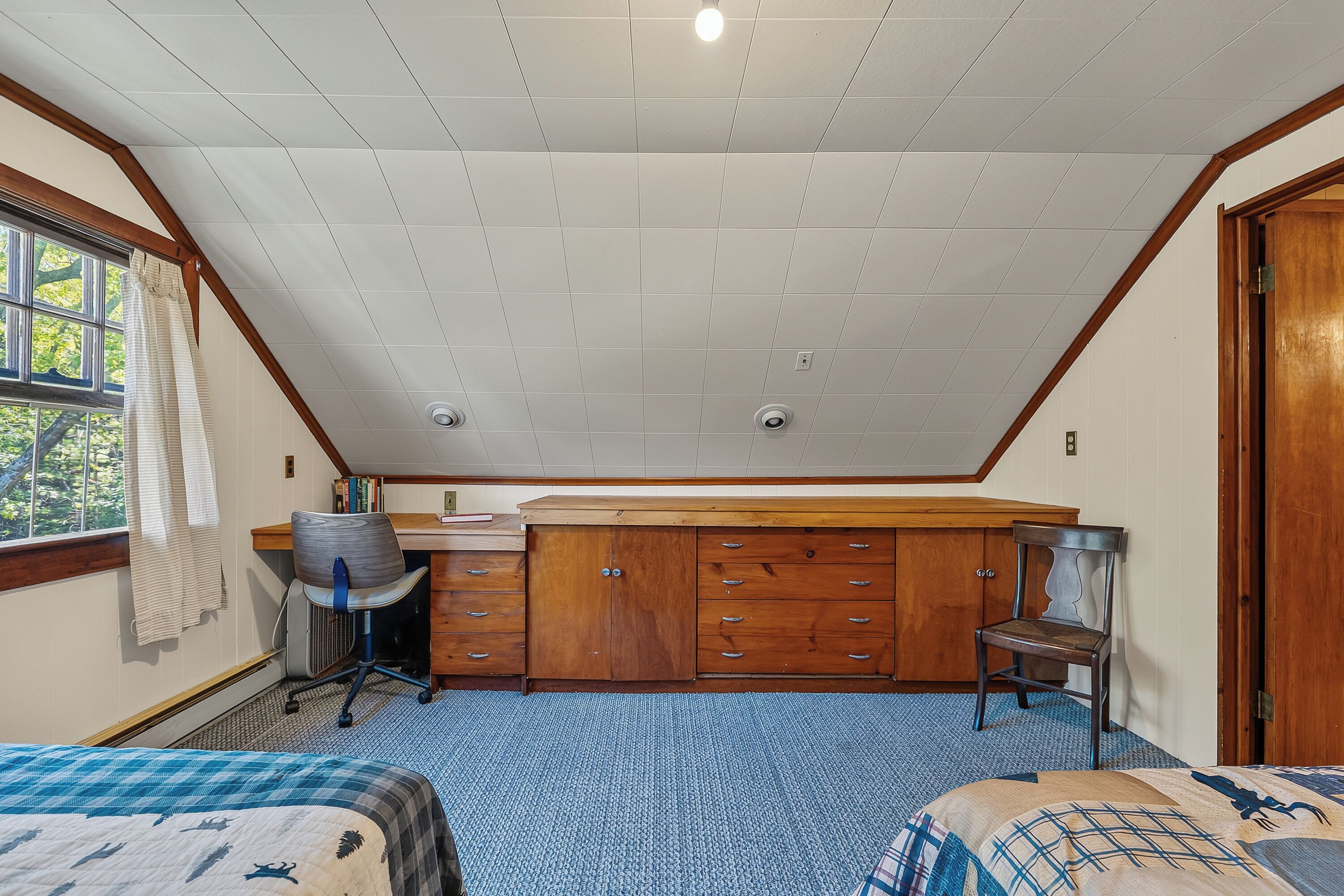A bedroom with a sloped ceiling, a desk, a chair, a wardrobe, and two beds.