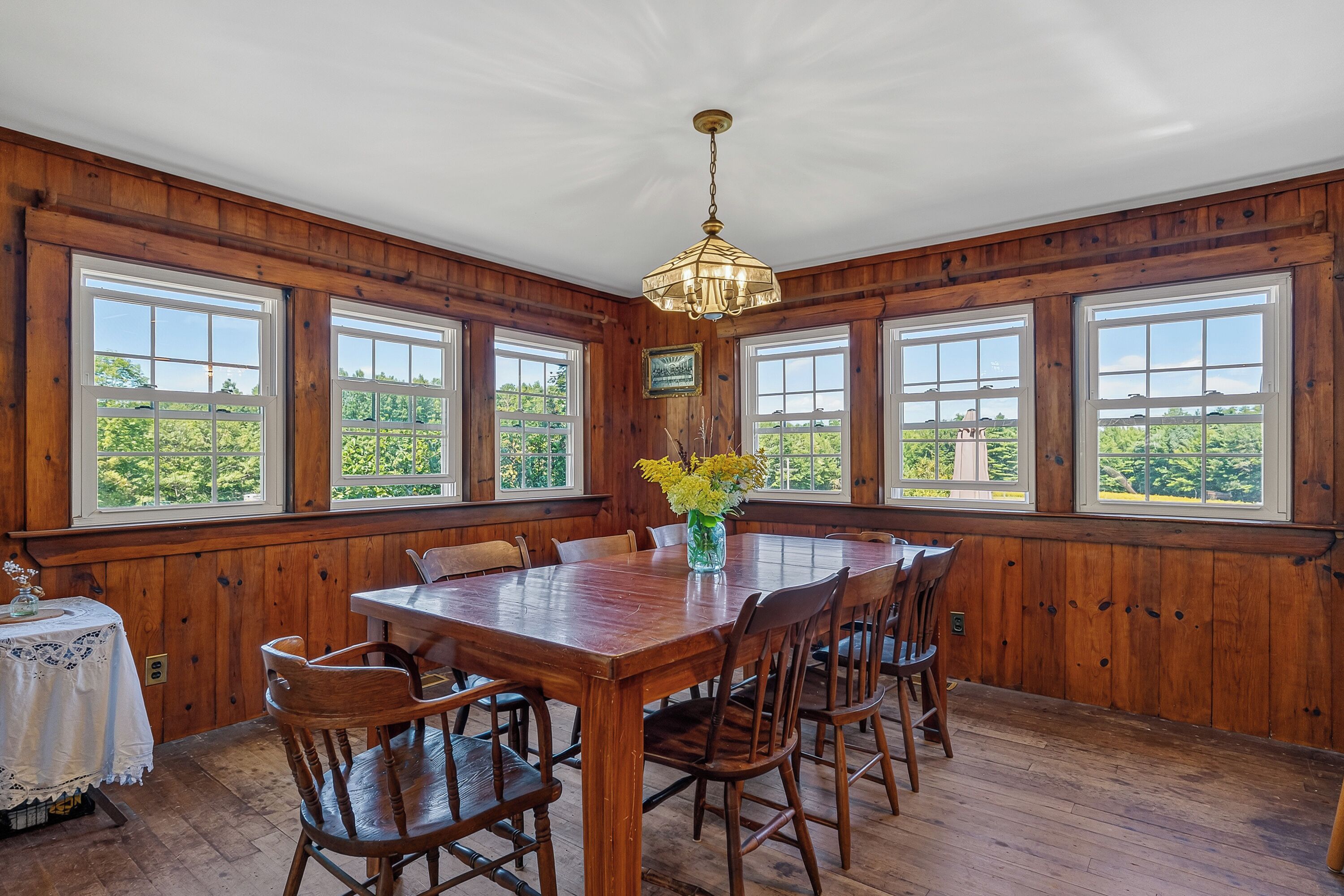 A dining room with a wooden table and chairs, large windows, and a chandelier.