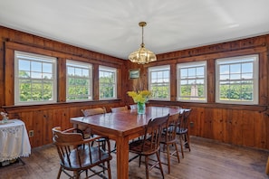 A dining room with a wooden table and chairs, large windows, and a chandelier. - 5-bedroom Lodge on 10 acres great for family reunions, retreats & weddings (Otisfield)