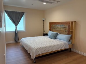 Ein Schlafzimmer mit einem Holzbettgestell, einer gemusterten Bettdecke, einem Nachttisch mit Lampe und einem Fenster mit Vorhängen.