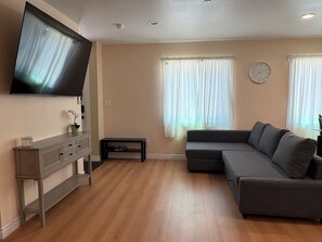 Ein Wohnzimmer mit einem an der Wand befestigten Flachbildfernseher, einem grauen Sofa, einem kleinen Tisch mit einem Paar Schuhen und einer Wanduhr.