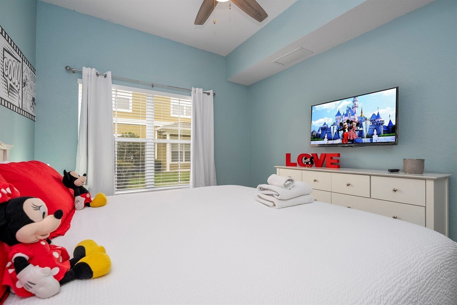 A bedroom with a large bed, a television mounted on the wall, a window with curtains, and a Mickey Mouse plush toy on the bed.