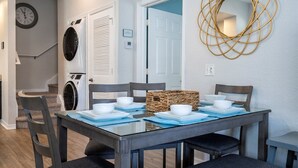 A dining area with a rectangular table set for four, surrounded by dark chairs. A woven basket is placed on the table, and a wall-mounted mirror with a gold frame is visible.