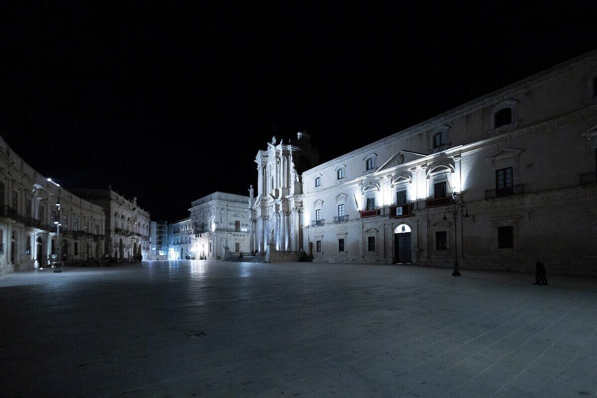 A large, open plaza with historic buildings, including a prominent church with a bell tower, under nighttime lighting.