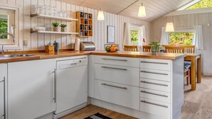 Eine moderne Küche mit weißen Schränken, Holz Arbeitsplatten und Pendelleuchten. Es gibt eine Essecke mit Tisch und Stühlen, und ein Fenster mit Blick auf Grünflächen.