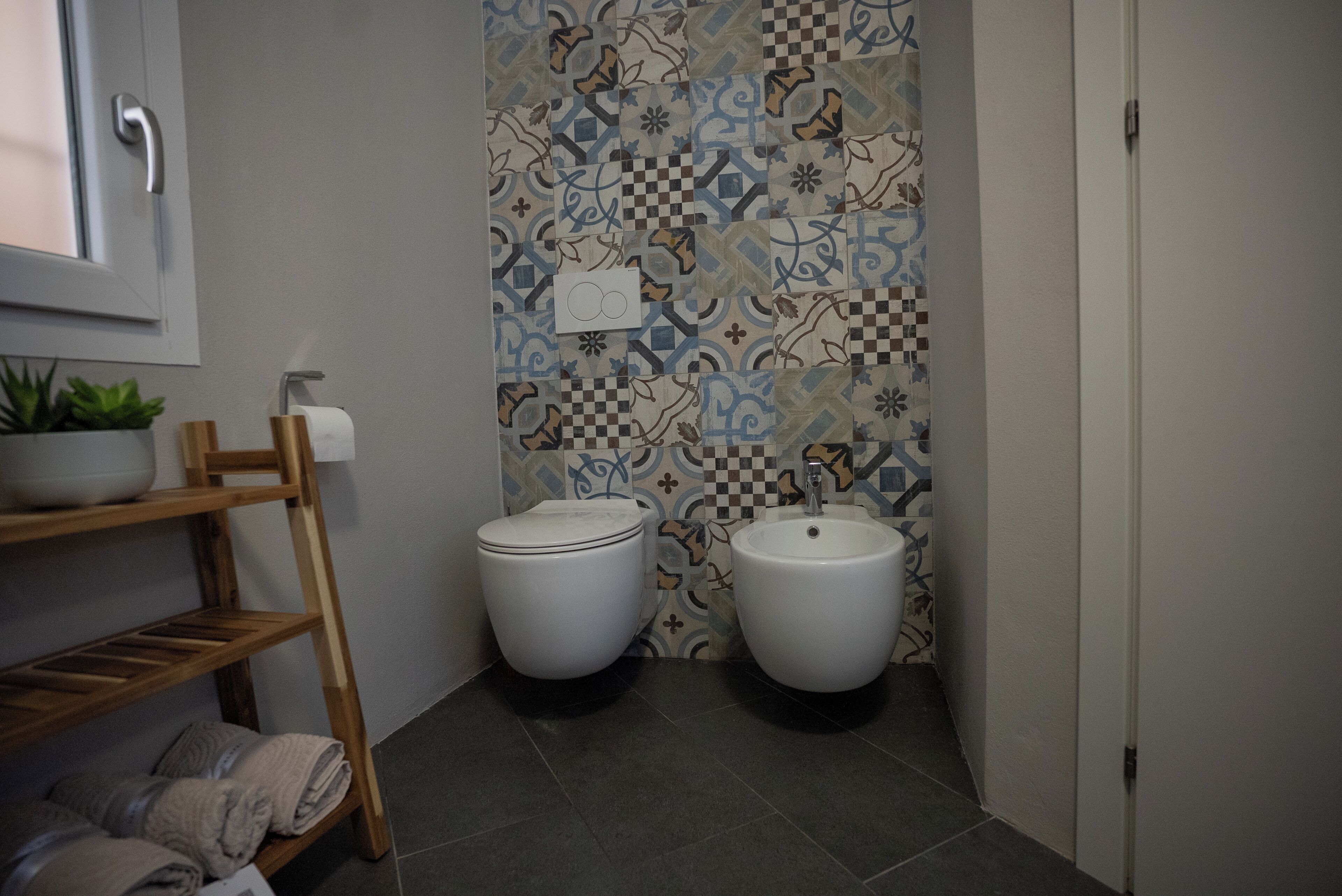 A bathroom with two toilets, a sink, a shelf with towels, and a patterned wall.