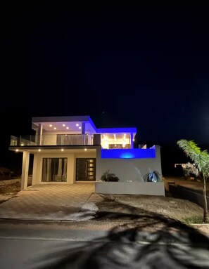 A modern two-story house with a balcony, large windows, and a well-lit entrance. - Ocean View Luxury Villa • Heated Pool • 5-Min Walk to Jeremi Beach (Lagun)